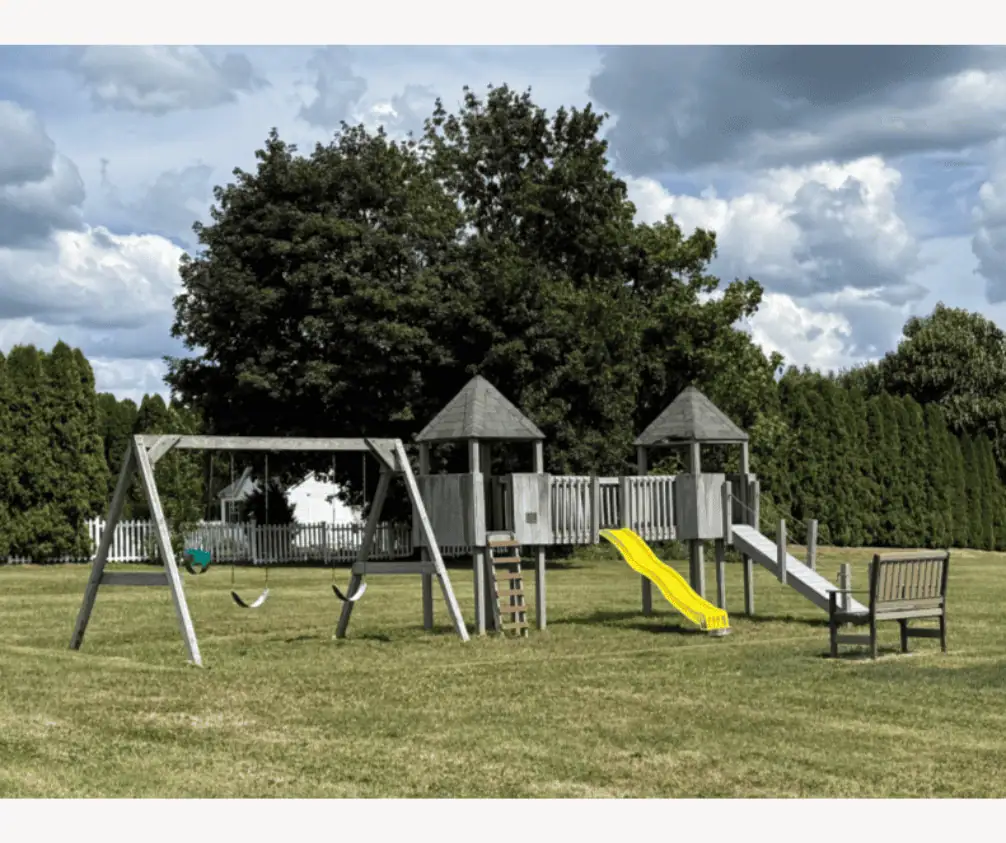A grey wooden children's play area in a grassy field with green trees behind it under a cloudy sky. The play area has two swings, two elevated huts with a bridge between them and a bright yellow slide. There is a bench facing the play area. A grey wooden children's play area in a grassy field with green trees behind it under a cloudy sky. The play area has two swings, two elevated huts with a bridge between them and a bright yellow slide. There is a bench facing the play area.