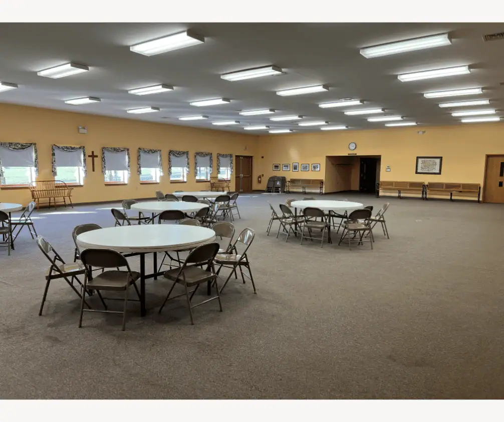 A photo of a large, bright, cheerful room used for large gatherings. The walls are a warm peach color and the carpeting is grey. Seven windows are visible and there are five white round tables set up with folding chairs around them. A photo of a large, bright, cheerful room used for large gatherings. The walls are a warm peach color and the carpeting is grey. Seven windows are visible and there are five white round tables set up with folding chairs around them.