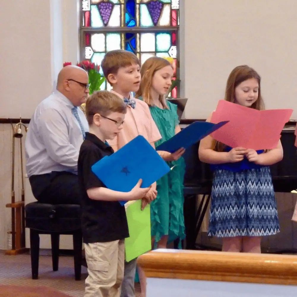 A photo taken in Schoeneck's sanctuary of the Junior Choir from a few years ago. A man plays the black grand piano and four nicely dressed boys and girls hold brightly colored folders while they sing.