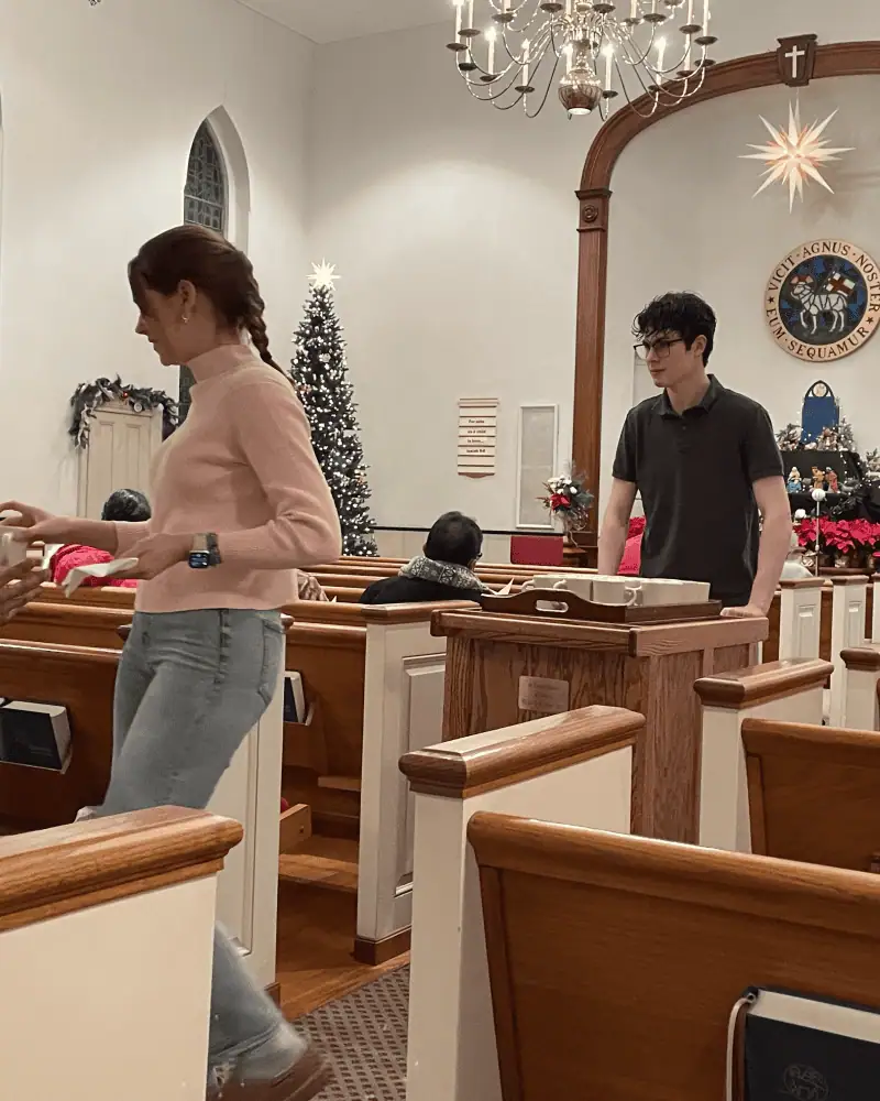 Two Schoeneckian youth - one boy and one girl - serve the lovefeast during the New Year's Eve service. The young woman is handing a coffee mug to a congregation member while the young man pushes the lovefeast cart.