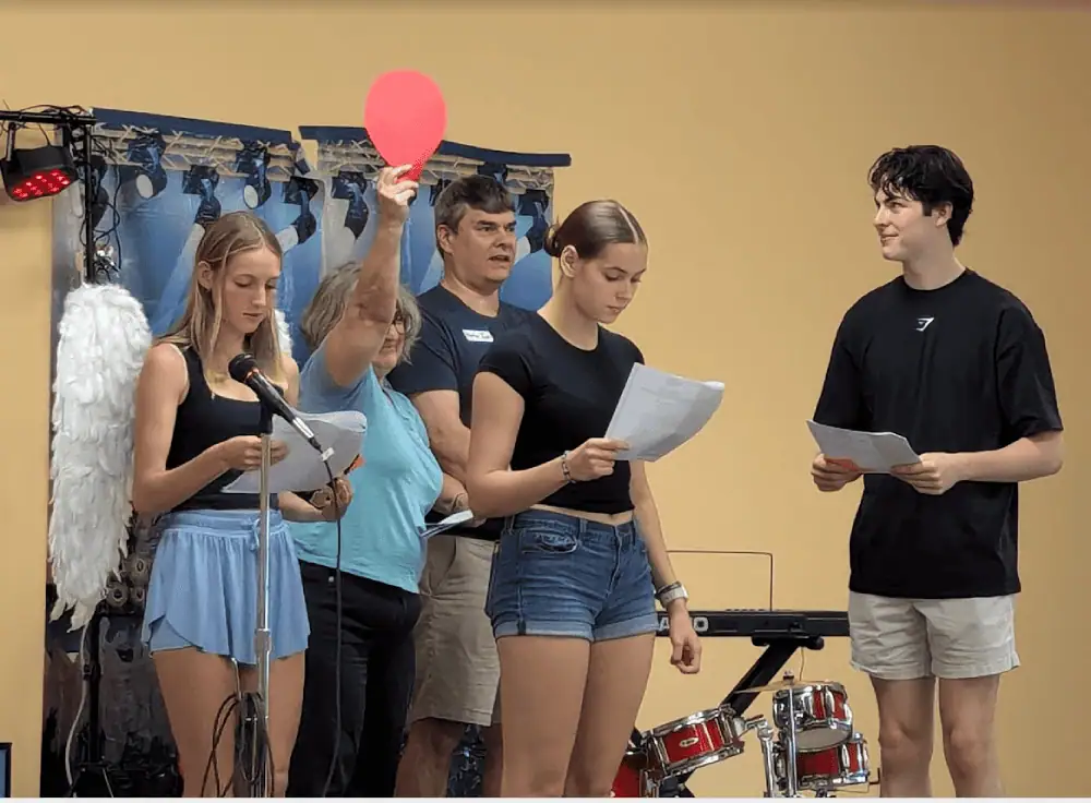 Three Schoeneckian youth (two girls and a boy) stand with Pastor Ian Edwards and Pastor Eileen Edwards on stage during VBS. One of the young ladies is wearing angel wings. Pastor Eileen is holding up a red paper balloon.