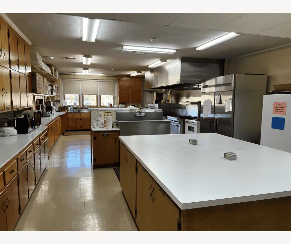 A photo showing the prep and serve area of the Church Kitchen. The floor is a tan linoleum. In the center of the picture is a large island with a white countertop and cabinets underneath. Behind the island is a large stainless steel sink. Along the left side of the photo is a long countertop with cabinets above and below stretching towards the prep and cook area of the kitchen with a three windows above the sink. On the right side of the photo are a large double-door industrial refrigerator and a separate white stand-up freezer. A photo showing the prep and serve area of the Church Kitchen. The floor is a tan linoleum. In the center of the picture is a large island with a white countertop and cabinets underneath. Behind the island is a large stainless steel sink. Along the left side of the photo is a long countertop with cabinets above and below stretching towards the prep and cook area of the kitchen with a three windows above the sink. On the right side of the photo are a large double-door industrial refrigerator and a separate white stand-up freezer.