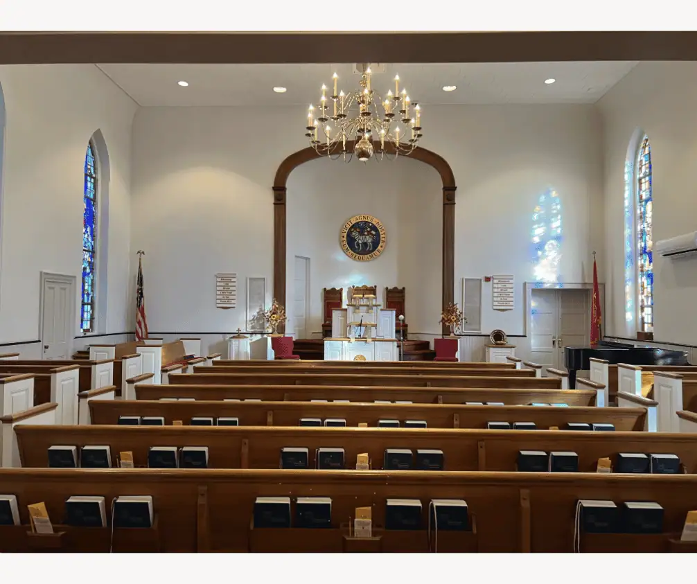 A photo of the Sanctuary taken from the back and looking towards the pulpit. There are several rows of pews visible, with blue hymnals in the pew racks. The pews are divided into three sections - two smaller sections on the outsides with a larger center section. There are two aisles. The pews are a warm medium wood with white ends. There are two chandeliers. Two stained glass windows are visible, one on each side of the picture. At the top of the picture is the two-level pulpit. The lower level has a white communion table and two burgundy chairs. The upper level has a white lectern and three chairs. Schoeneck's Moravian Seal is visible on the back wall of the pulpit. A photo of the Sanctuary taken from the back and looking towards the pulpit. There are several rows of pews visible, with blue hymnals in the pew racks. The pews are divided into three sections - two smaller sections on the outsides with a larger center section. There are two aisles. The pews are a warm medium wood with white ends. There are two chandeliers. Two stained glass windows are visible, one on each side of the picture. At the top of the picture is the two-level pulpit. The lower level has a white communion table and two burgundy chairs. The upper level has a white lectern and three chairs. Schoeneck's Moravian Seal is visible on the back wall of the pulpit.