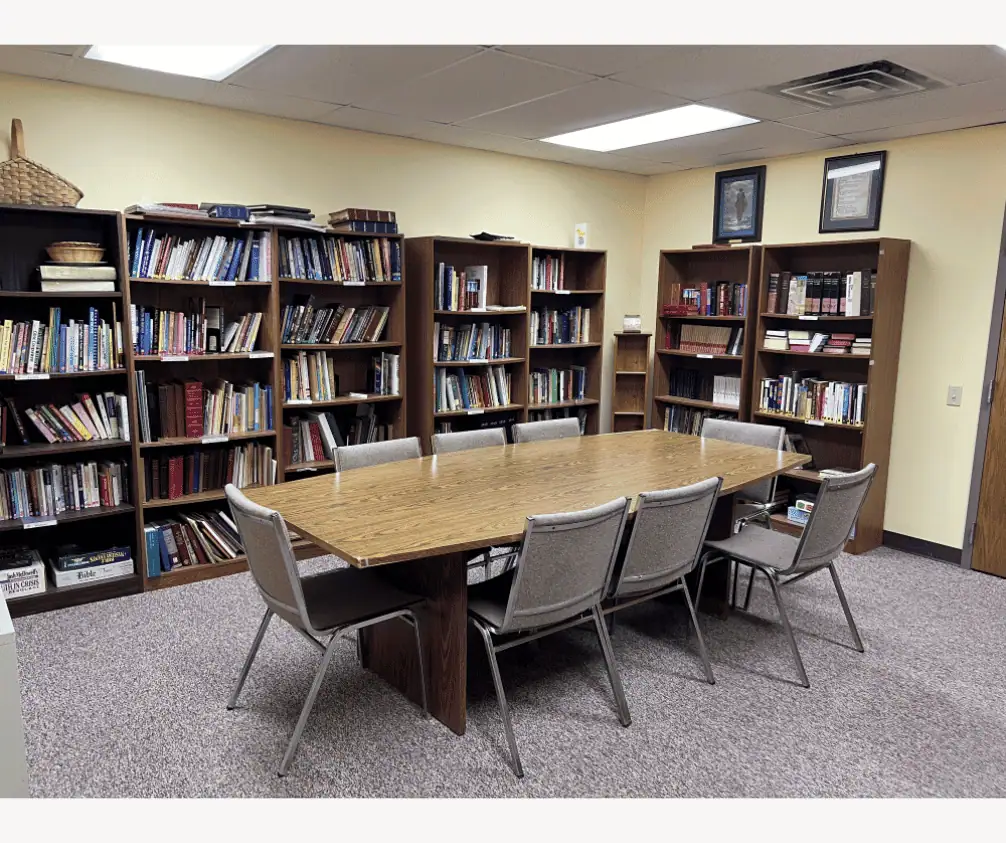 A photo of a grey carpeted room with light yellow walls. There is a brown table in the middle with eight grey chairs surrounding it. The walls are lined with dark brown bookshelves filled with books, videos, and other resources. A photo of a grey carpeted room with light yellow walls. There is a brown table in the middle with eight grey chairs surrounding it. The walls are lined with dark brown bookshelves filled with books, videos, and other resources.