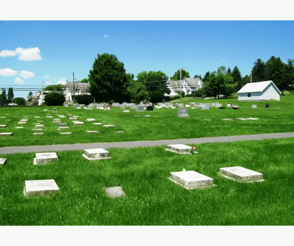 A color picture of the Schoeneck cemetery, known as God's Acre. The sky is a bright sunny blue and the grass is a verdant green. There are flat white markers at the bottom of the picture. Just above the markers is a gravel road. At the top of the image are more flat white markers. A color picture of the Schoeneck cemetery, known as God's Acre. The sky is a bright sunny blue and the grass is a verdant green. There are flat white markers at the bottom of the picture. Just above the markers is a gravel road. At the top of the image are more flat white markers.