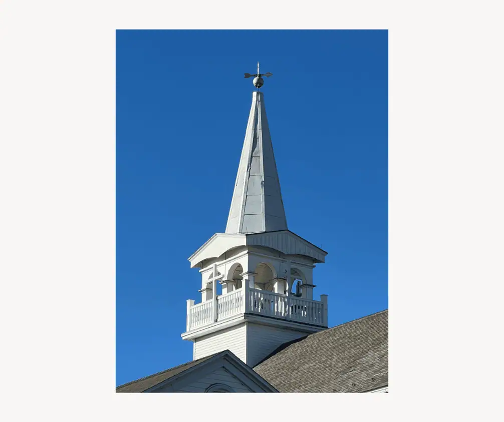 The picture shows the white bell tower and steeple of the church against a blue sky. The belfry is square with a railing around it. A white cross is visible on the side of the belfry. The steeple rises into the sky with a weathervane on top. The picture shows the white bell tower and steeple of the church against a blue sky. The belfry is square with a railing around it. A white cross is visible on the side of the belfry. The steeple rises into the sky with a weathervane on top.
