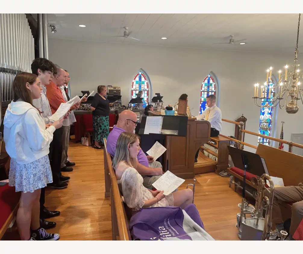 A view of the balcony during worship. In the foreground, there is a row of people standing in front of the organ pipes singing from a lovefeast ode, and a couple people sitting on a pew in front of them. Some black music stands are visible, as well as several brass instruments. Director of Music Ministries, Ryan Morrow, sits at the organ console. In the background of the picture, several people stand and sing in front of handbell tables. A view of the balcony during worship. In the foreground, there is a row of people standing in front of the organ pipes singing from a lovefeast ode, and a couple people sitting on a pew in front of them. Some black music stands are visible, as well as several brass instruments. Director of Music Ministries, Ryan Morrow, sits at the organ console. In the background of the picture, several people stand and sing in front of handbell tables.