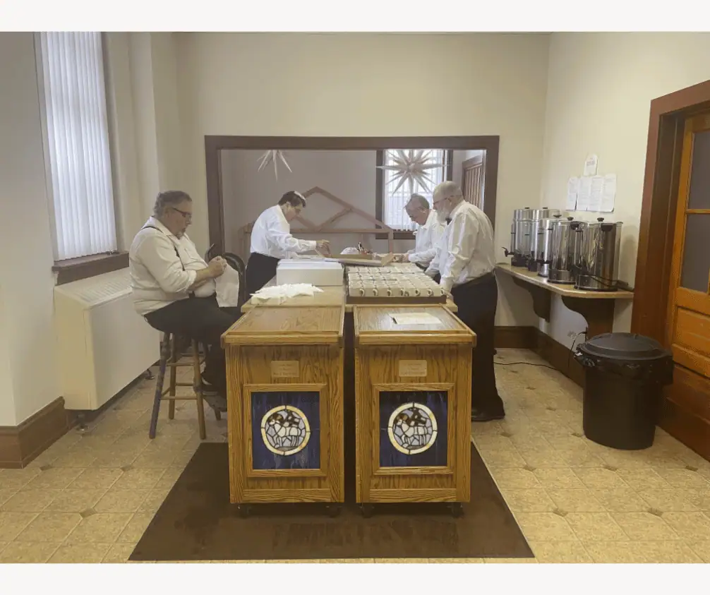 The photo shows a room with updated linoleum on the floor, a large window on the left, and a smaller room in the back with a white Moravian star hanging from the ceiling. In the center of the main room are two wooden carts with stained glass Angus Dei images. Behind the carts are a large island. Two men and two women are gathered around the island. Two of the people are wrapping pieces of sugar cake in white napkins. Two of the people are readying white coffee mugs onto trays. Along the right wall is a shelf with five large coffee urns. The photo shows a room with updated linoleum on the floor, a large window on the left, and a smaller room in the back with a white Moravian star hanging from the ceiling. In the center of the main room are two wooden carts with stained glass Angus Dei images. Behind the carts are a large island. Two men and two women are gathered around the island. Two of the people are wrapping pieces of sugar cake in white napkins. Two of the people are readying white coffee mugs onto trays. Along the right wall is a shelf with five large coffee urns.