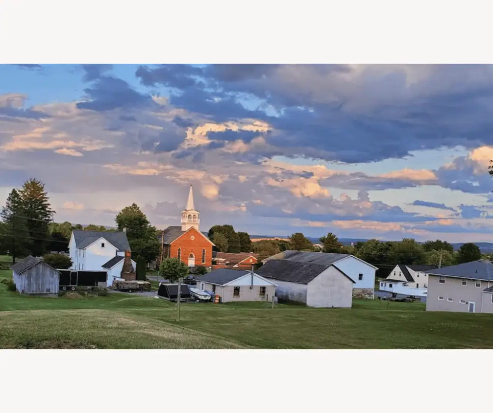 A picture of Schoeneck taken at sunset from a nearby neighborhood. It is sunset, and the sky is filed with clouds with golden and pink tinged edges. The church building is visible with just one of the peaked roofs of the Fellowship Hall. In front of the church are the buildings of the neighborhood, a white farmhouse and several out-buildings including a grey barn and a cement garage. In the foreground of the picture is a grassy field. A picture of Schoeneck taken at sunset from a nearby neighborhood. It is sunset, and the sky is filed with clouds with golden and pink tinged edges. The church building is visible with just one of the peaked roofs of the Fellowship Hall. In front of the church are the buildings of the neighborhood, a white farmhouse and several out-buildings including a grey barn and a cement garage. In the foreground of the picture is a grassy field.