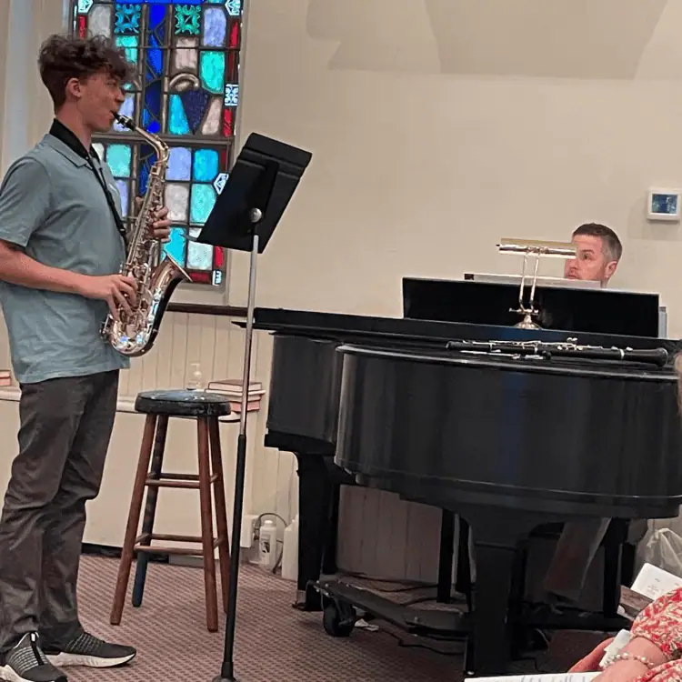 A young man plays the saxophone while Director of Music Ministries plays the black grand piano in Schoeneck's sanctuary.
