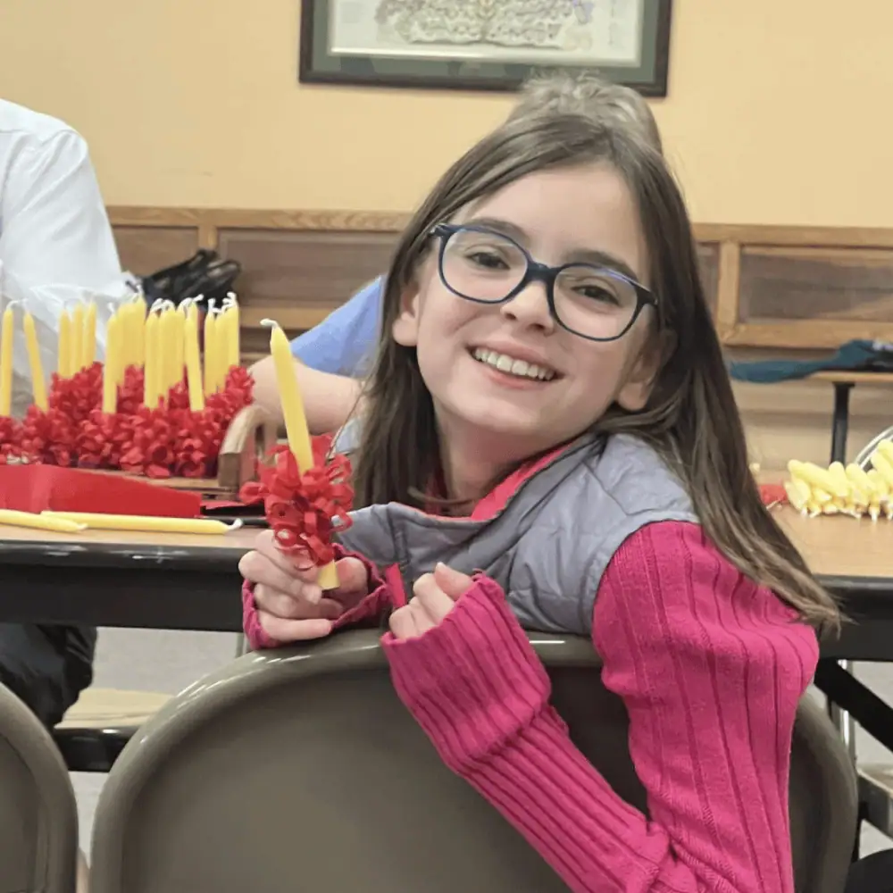 A pre-teen girl with a pretty smile leans over the back of a folding chair in Schoeneck's Fellowship Hall, proudly holding a beeswax candle that she just finished wrapping with a red frill.
