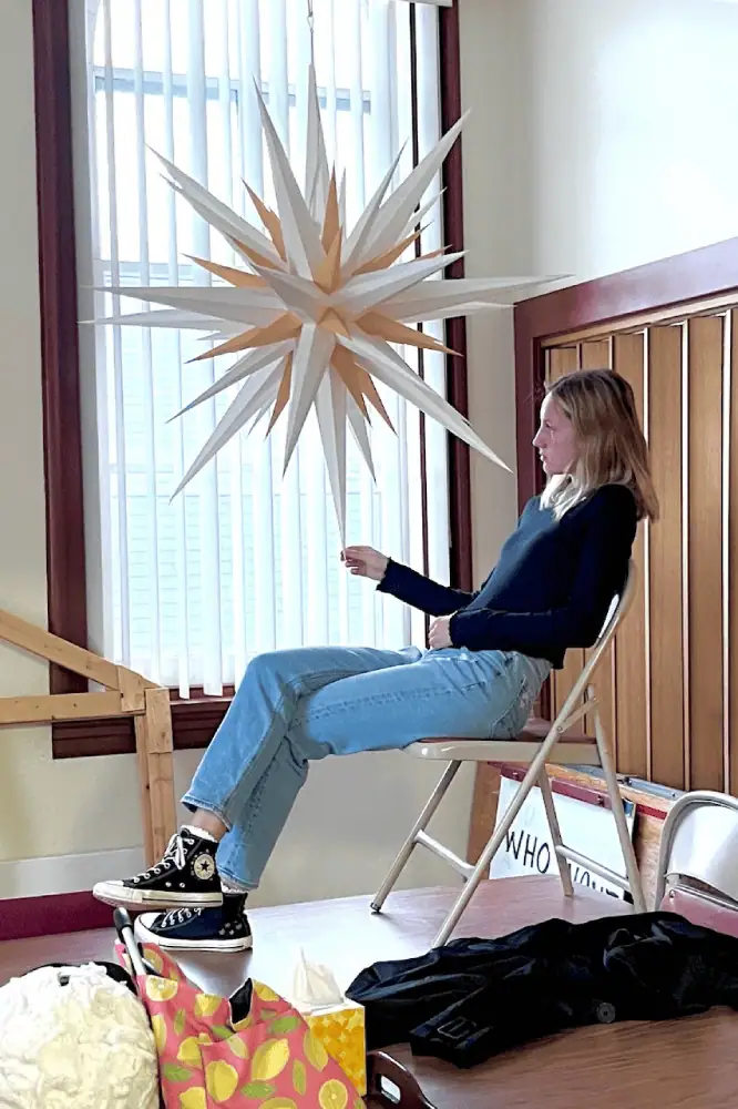 A young woman sits on a folding chair on top of a folding table in the Diener's Kitchen. Schoeneck's large Moravian Star hangs from the ceiling. The young lady is holding with bottom point of the star with a contemplative look on her face.