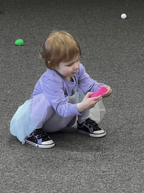 A young girl wearing a purple jumpsuit with a rainbow tutu and black sneakers squats in the Fellowship Hall, holding a bright pink plastic Easter egg.