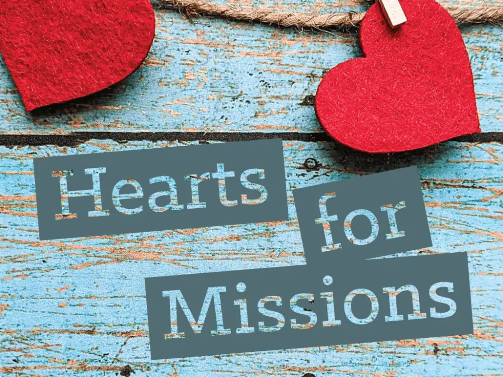 A clip-art image of a close-up of two rough light aquamarine wooden slats with two red felt hearts hung across the top with crafting string. Stenciled on the wood are the words Hearts for Missions.