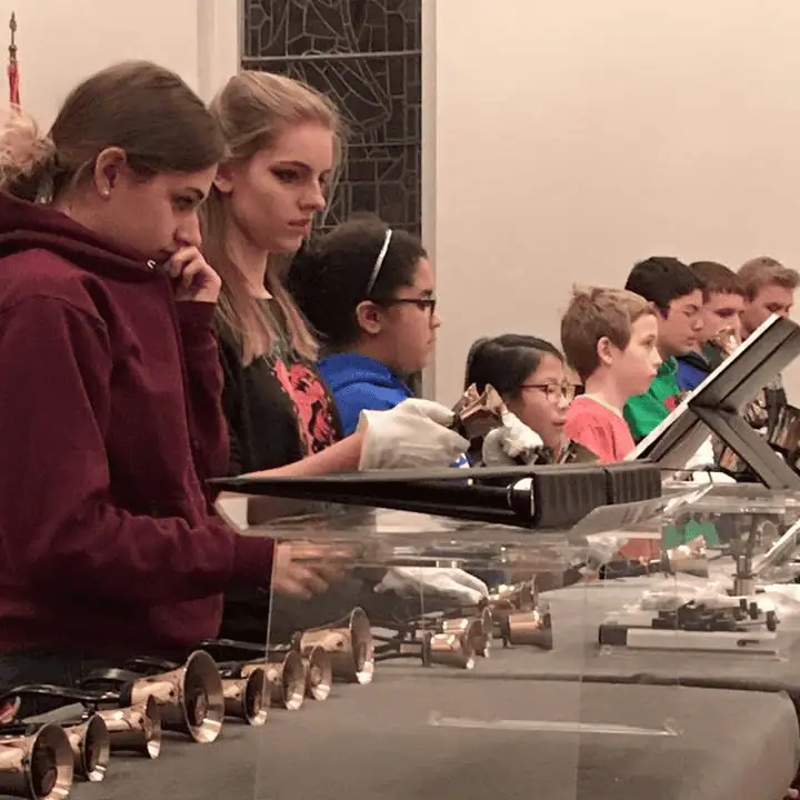 A group of young people stand behind several bell tables preparing for rehearsal. Some of the kids are putting white gloves on. Some of them are looking at the music, and some have already picked up their bells and are ready to play.