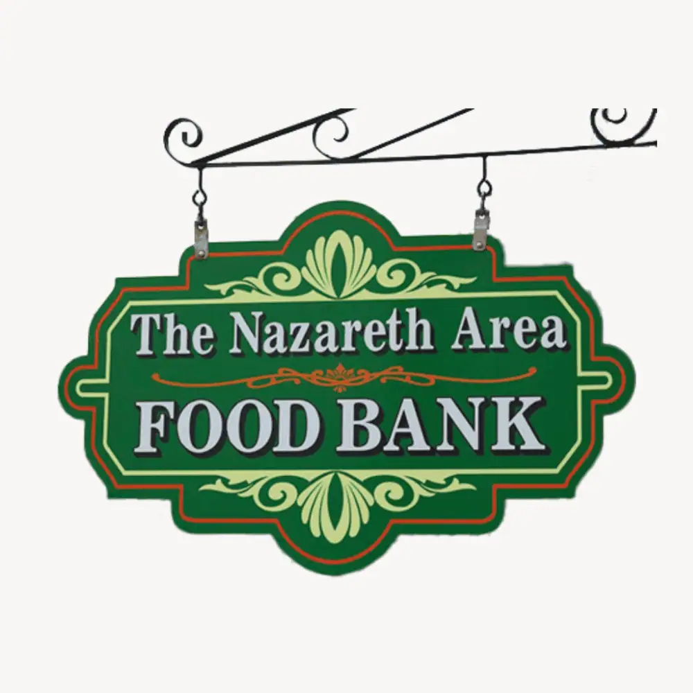 A photo of the Nazareth Area Food Bank sign, There is a black wrought iron bracket holds the sign, which is dark green with pale lime green and orange decorative accents. The words "The Nazareth Area Food Bank" are a classic serif font in white with a black drop shadow.