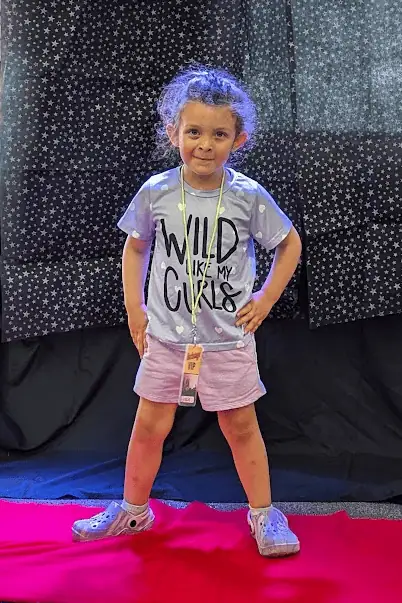 An older toddler stands in front of a shiny black backdrop with her hands on her hips as she poses for a Red Carpet picture.