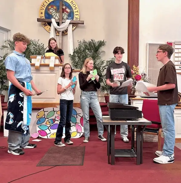 Six youth present their annual Youth Worship Service for the Schoeneck congregation. One youth is in the upper pulpit. One youth is wearing a beach towel over his clothes. Three youth have brightly colored cardboard fish in their hands, and one youth stands beside a portable grill.