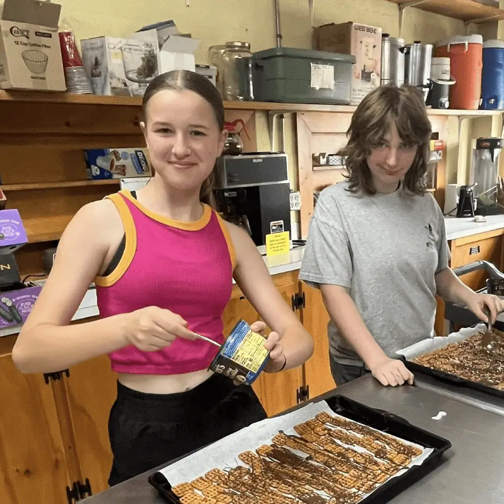 Two teen girls drizzle chocolate over trays of square pretzels in Schoeneck's kitchen.