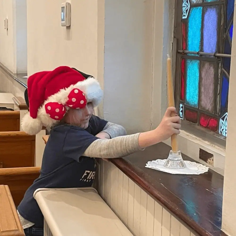 A pre-teen girl wearing a Santa hat places a large beeswax candle in a candleholder on one of the windowsills in front of a stained glass window in Schoeneck's sanctuary.
