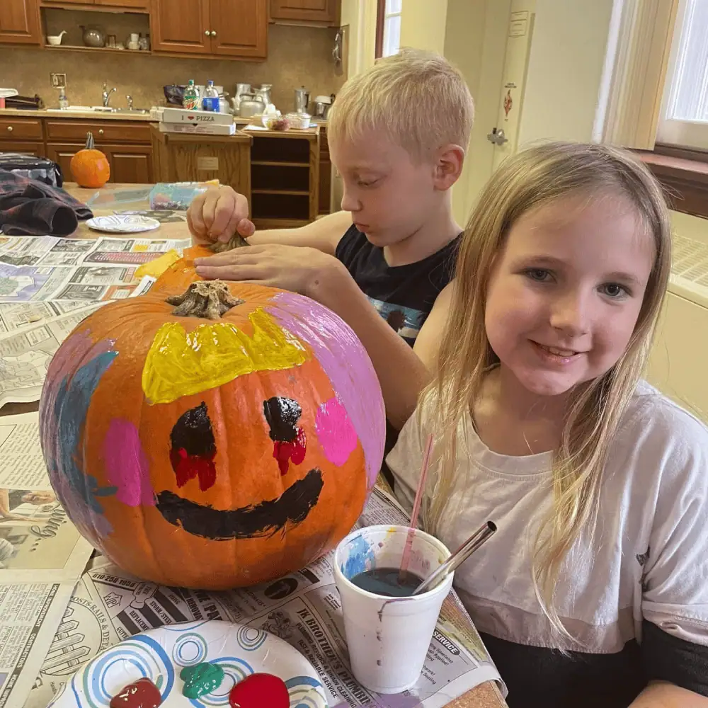 A boy and a girl are in the Diener's Kitchen at Schoeneck. The boy is putting the top back on his carved pumpkin, and the girl is posing with her painted pumpkin.