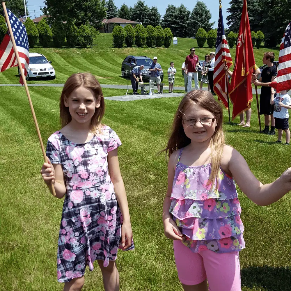 Two preteen girls wave small American flags on sticks as they wait for the annual Memorial Day service in God's Acre to begin. Behind them, Schoeneck's Brass Choir is setting up, and some Boy Scouts prepare an honor guard with two large American flags.