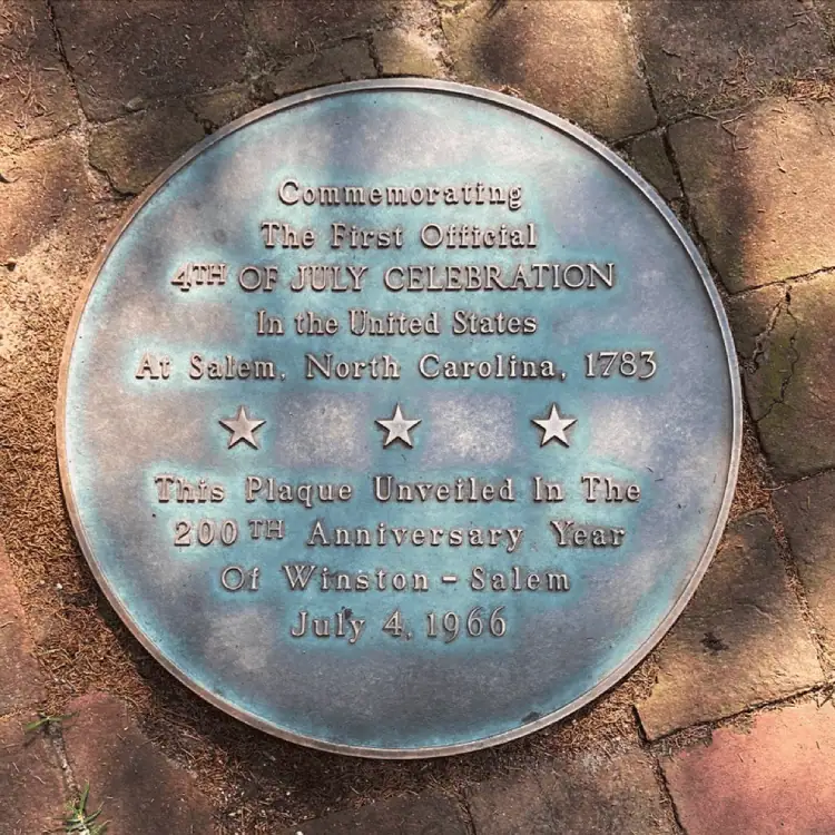 A close-up photo of a copper and green plaque in a red brick sidewalk. The text on the plaque reads: Commemorating the first official 4th of July celebration in the United States at Salem, North Carolina, 1783. There are three stars in a row and then the words continue with: This plaque unveiled in the 200tyh anniversary year of Winston-Salem, July 4, 1966.