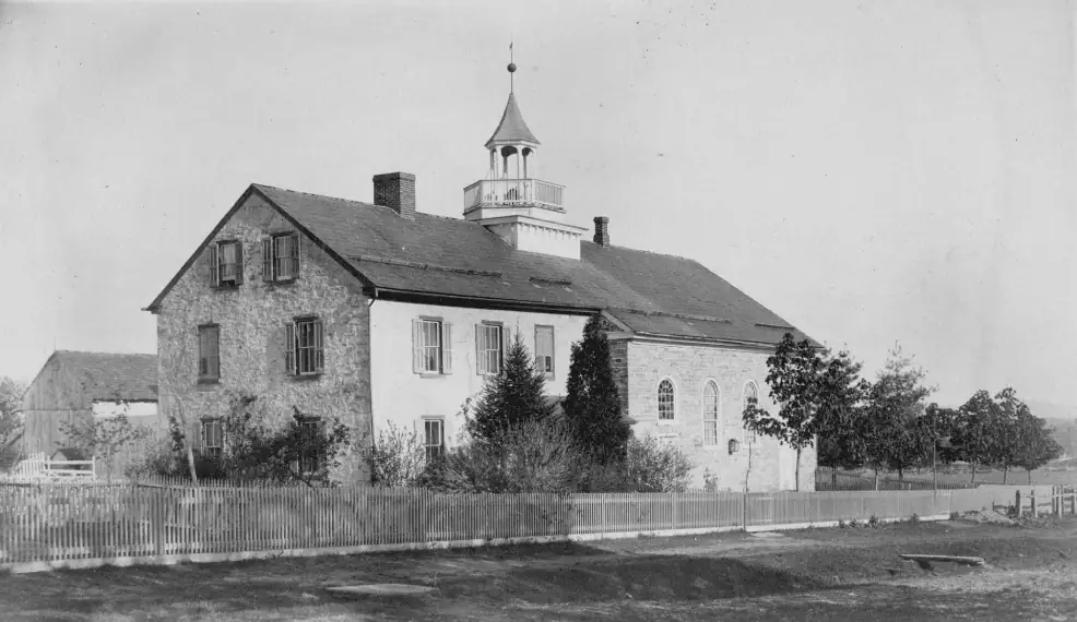 A black and white photo of the 1793 Schoeneck parsonage and church from around 1887. The two buildings are connected. The parsonage is on the left and is made of stone. There are windows on three floors of the parsonage - two small windows in the attic space, two windows on the side on the first and second floors, and three windows on the front of both floors. A window on the first floor is visible, but another window and the porch covered door are obscured behind trees and bushes. The church is on the right side of the parsonage and is also built of stone. There are two doors, one slightly obscured by some bushes, one at each end of the building. Above each door is a small arched window. There are two longer arched windows in the middle, at the same level as the smaller windows. The church has a small bell tower, built with a balcony around it for the brass choir to play from. There is a picket fence in front of the church, along with more shrubbery and trees. Behind the church, part of a barn is visible.