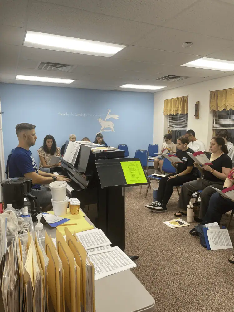 The Senior Choir rehearses in the Choir Room. Director of Music Ministries, Ryan Morrow, sits at a black upright piano while several men, women, and youth sit in blue chairs singing an anthem. The far wall of the room is painted light blue, with a cream image of the Angus Dei and the words: "Worthy is the Lamb for He was slain."