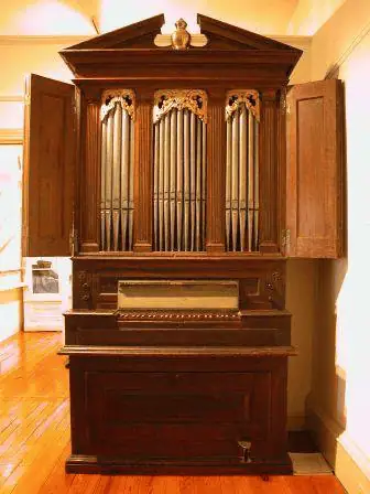 A photo of the 1776 Tannenberg organ at the Whitefield House. The organ is a dark cherry wood. The upper cabinet is open to show three sections of pipes. Underneath the cabinet is the organ keyboard with a bench.